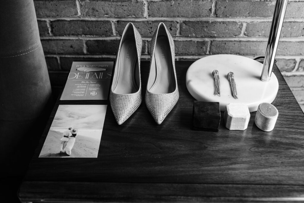Brides wedding details including her glittery shoes, wedding ring boxes, and invitations sit on wooden cabinet with brick background.
