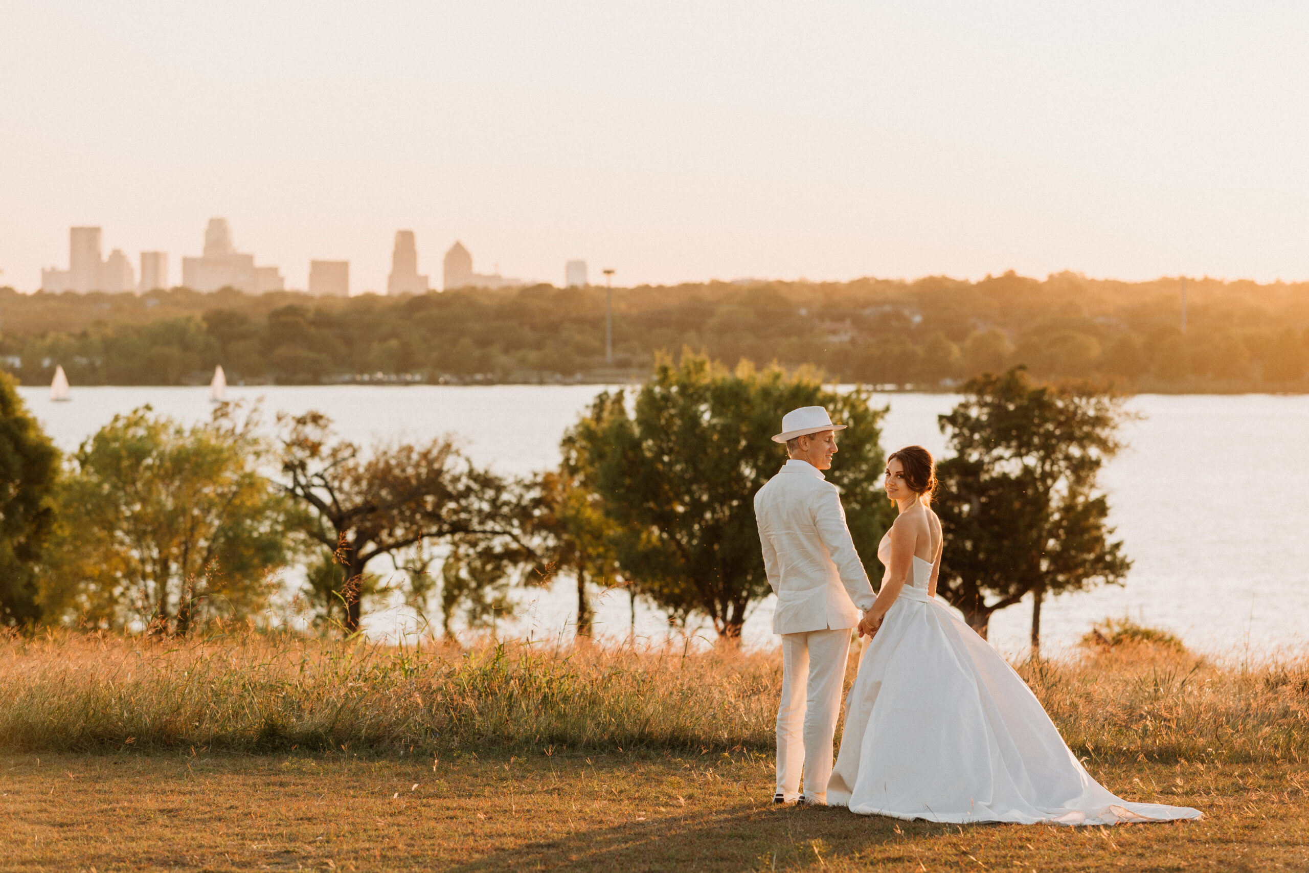 Bride in white dress stands holding hands with groom in white suit overlooking a scenic lake view with Dallas skyline in the background.
