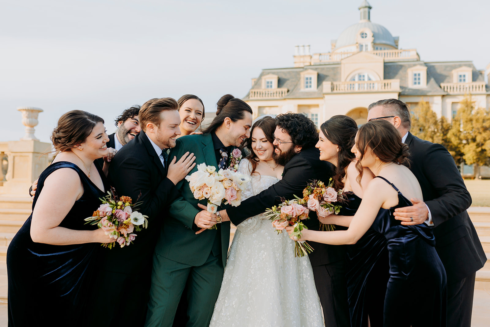 Bride and groom are surrounded and embraced by wedding party while standing in front of the extravagant facade of The Olana