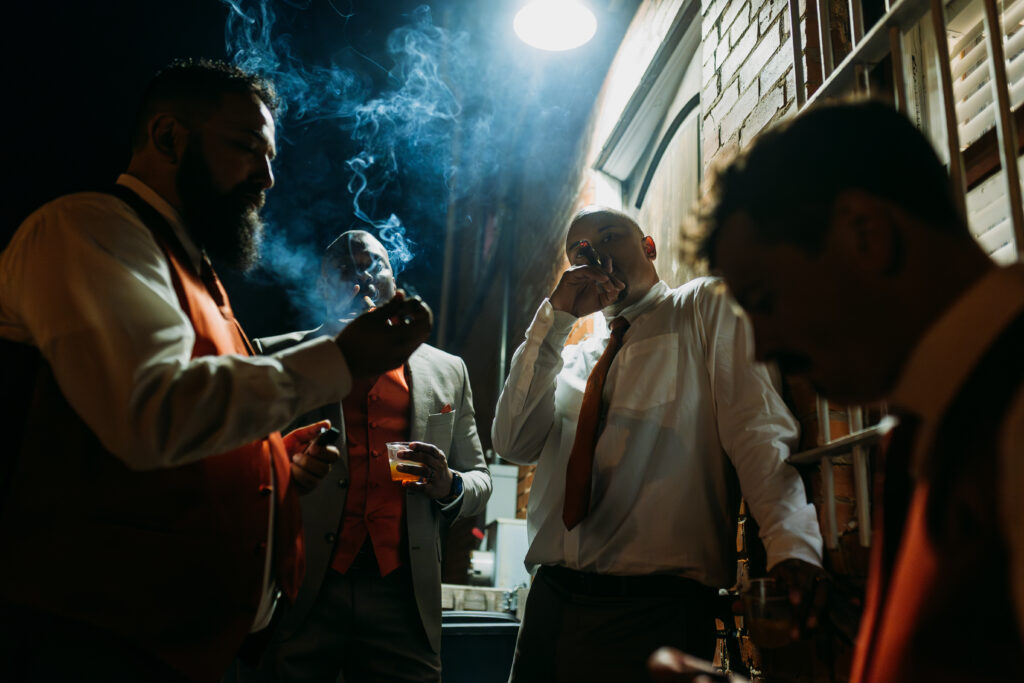 Groom and groom's party smoke cigars outside of wedding reception in stark light
