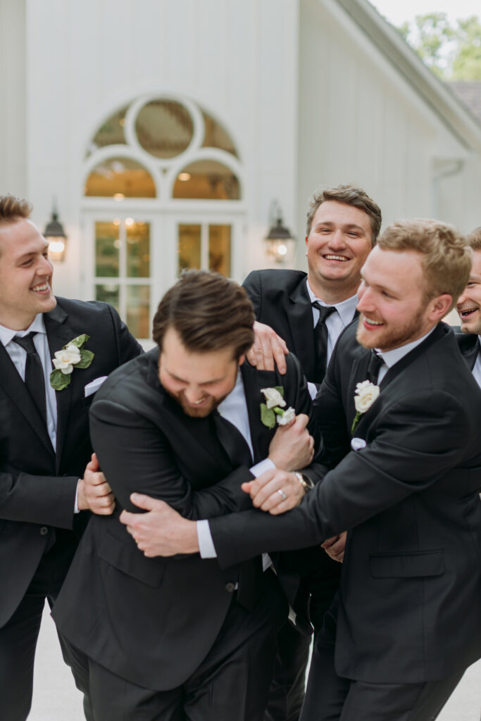 Grooms party tackles and jeers groom while laughing in front of chapel at The French Farmhouse
