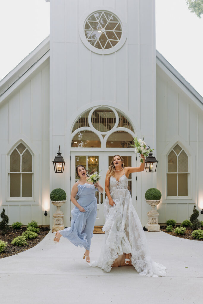 Bride and Bridesmaid jump and 
laugh while in front of the Chapel at The French Farmhouse in DFW
