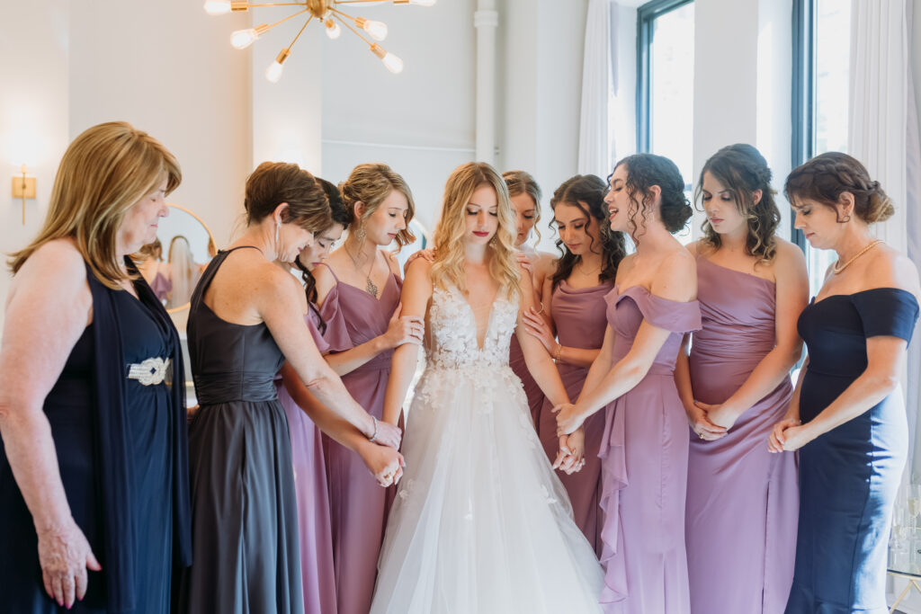 Bride is at center of her bridal party while they gather, hold hands, and pray
