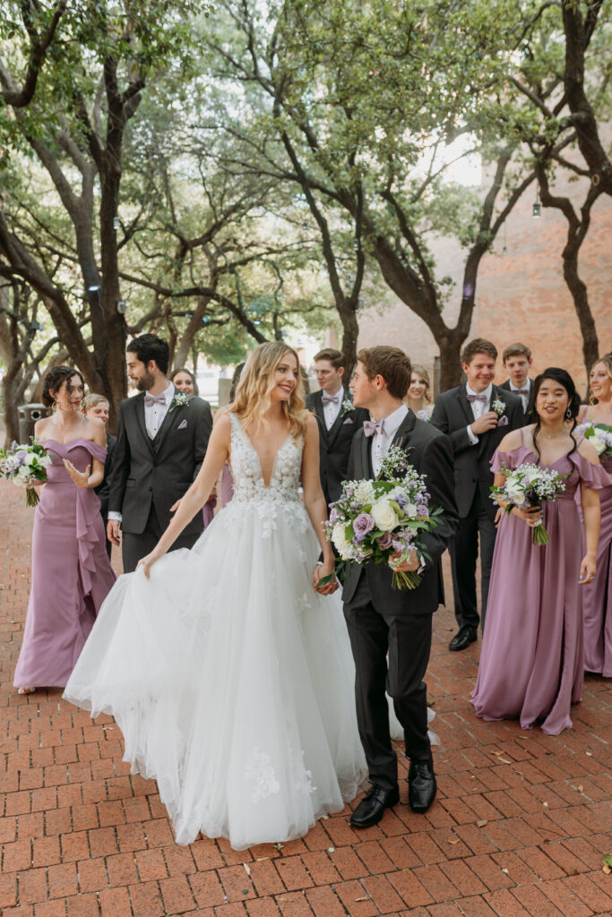 Bride and Groom walk hand in hand as wedding party follows loosely in park near The Century Hall in Downtown Fort Worth
